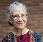 A woman smiling with gray hair and glasses. She wears a colorful striped garment with complex print and stands in front of a brick wall
