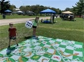 Two children playing on a giant board game with a giant die. They're outdoors on a sunny day with tents in the background.
