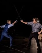 Two thespians dueling with swords on an outdoor stage at night.